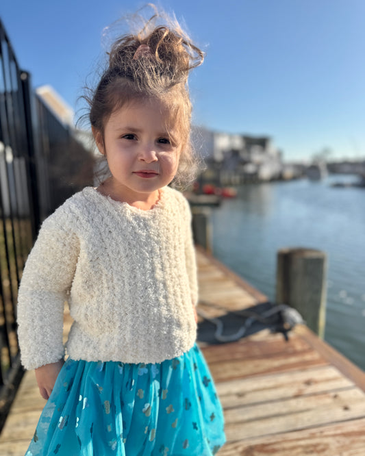 Child in a white sweater and blue dress standing on a dock with water and buildings in the background