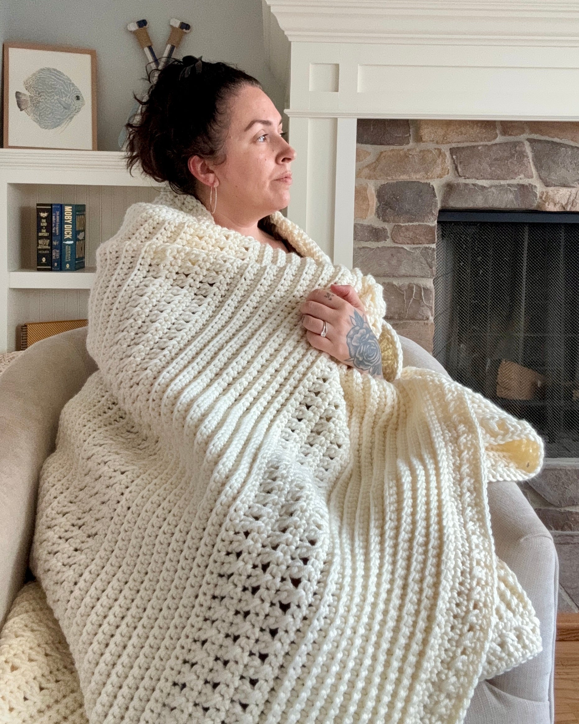 Woman wrapped in a large white crocheted blanket sitting in front of a fireplace.
