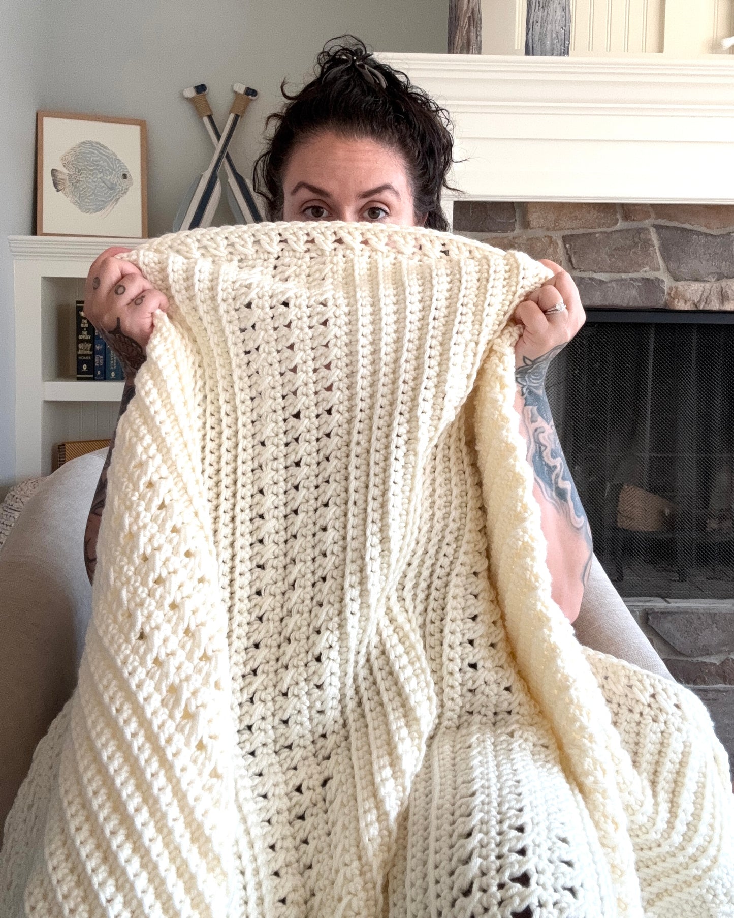 Person holding a large white crocheted blanket in front of them, with a fireplace and decor in the background.