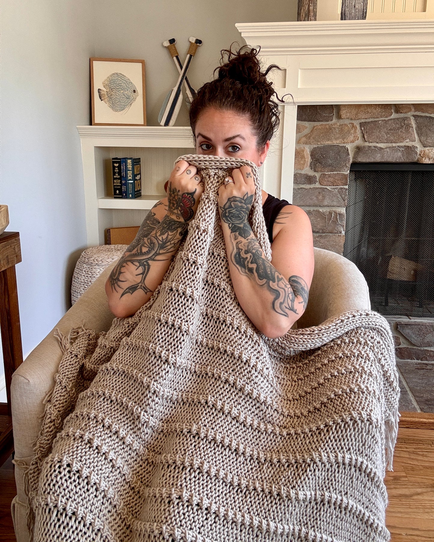 Woman with tattoos sitting on a couch, wrapped in a blanket in a cozy living room.