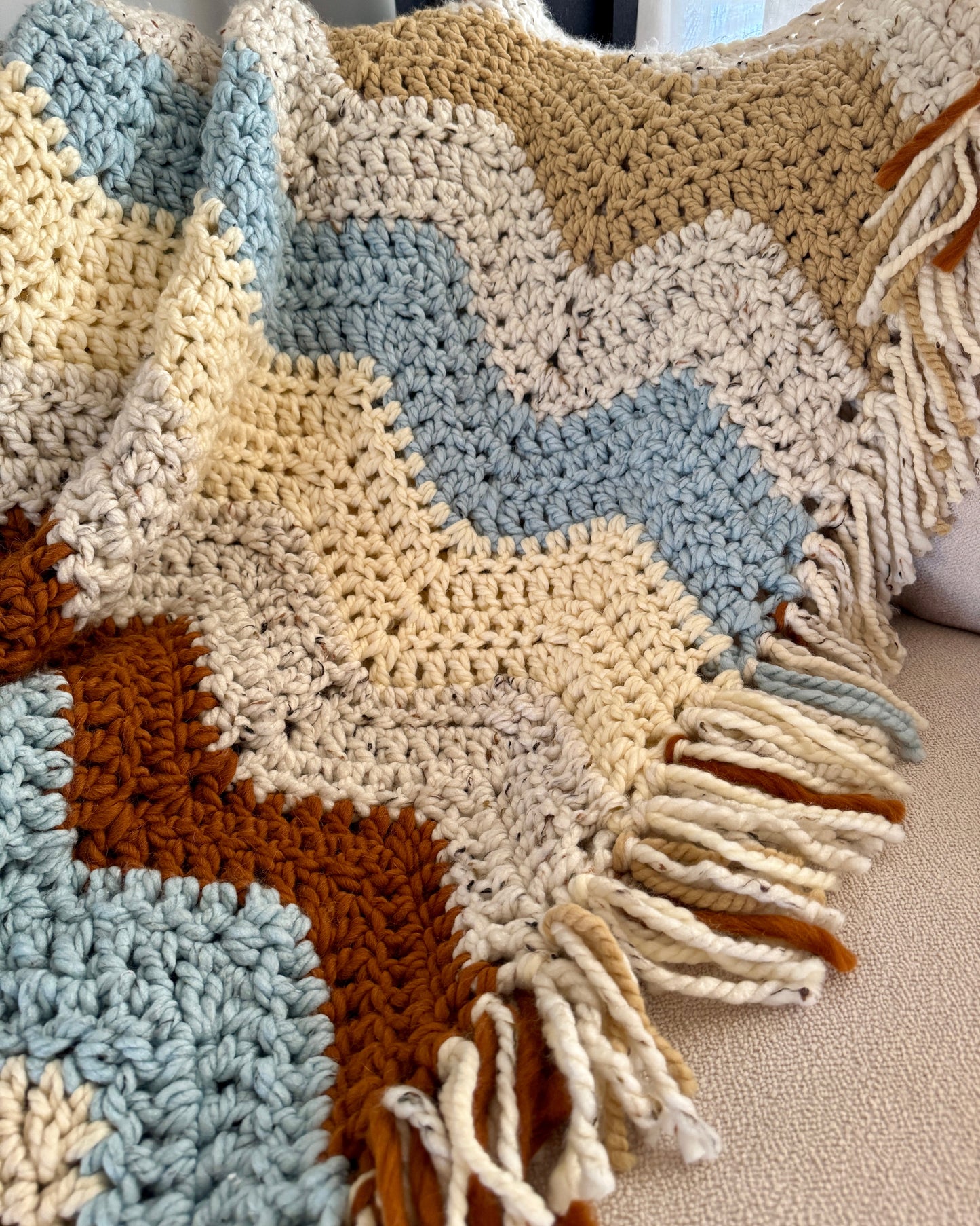 Crocheted blanket with a zigzag pattern in brown, beige, and blue on a neutral background.