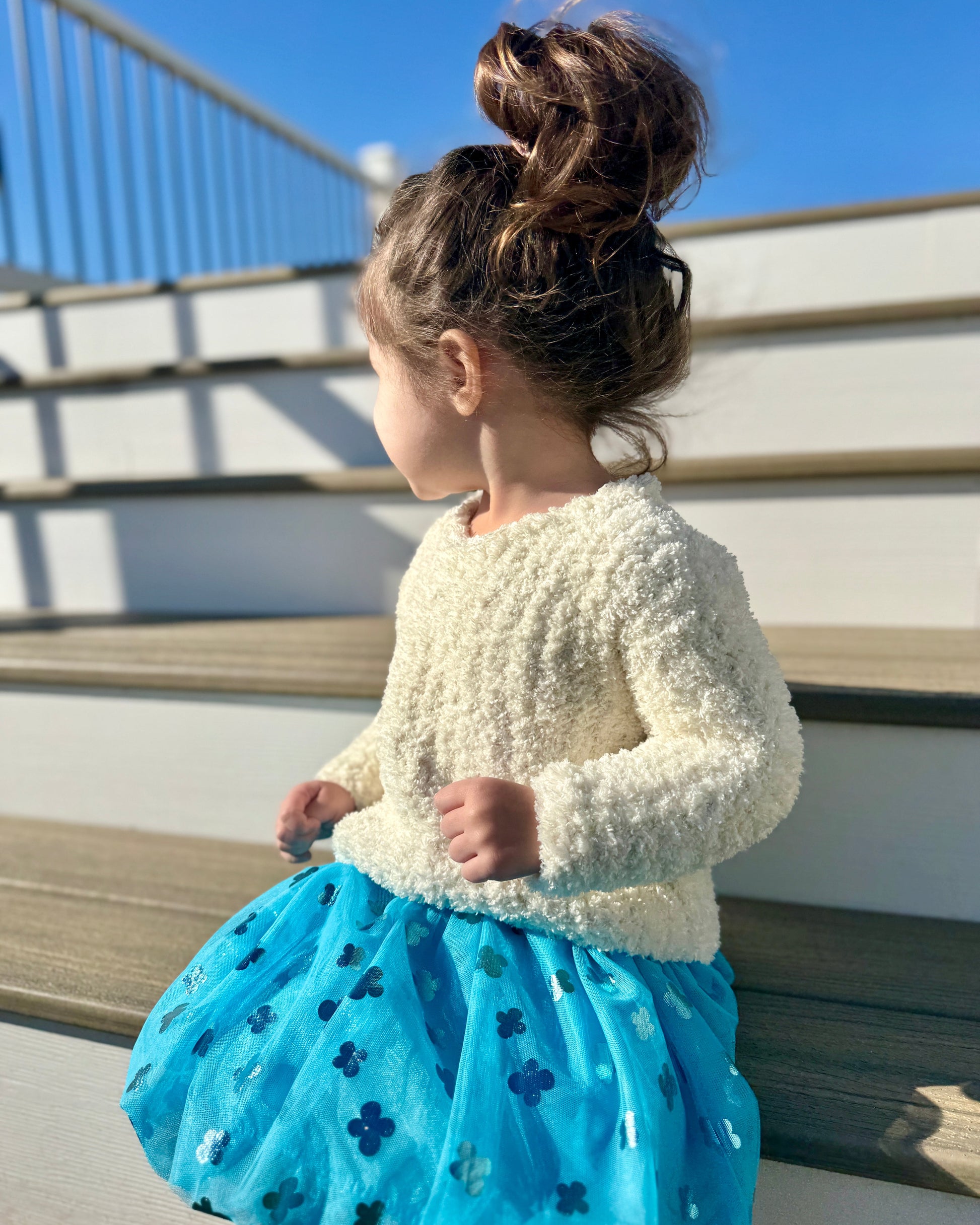 Child wearing a white sweater and blue dress with star patterns, standing on a wooden deck.