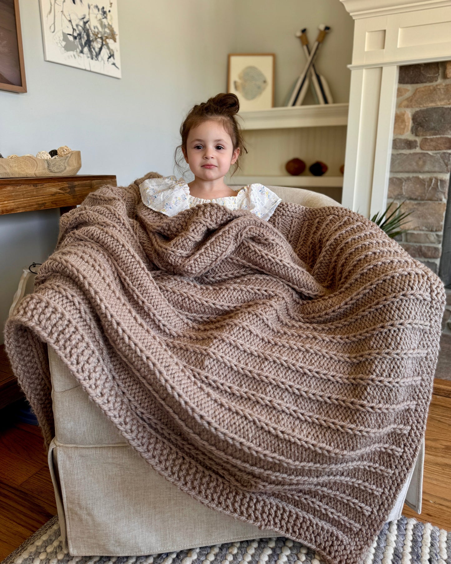 Child sitting on a couch with a large brown knitted blanket, in a cozy living room setting.