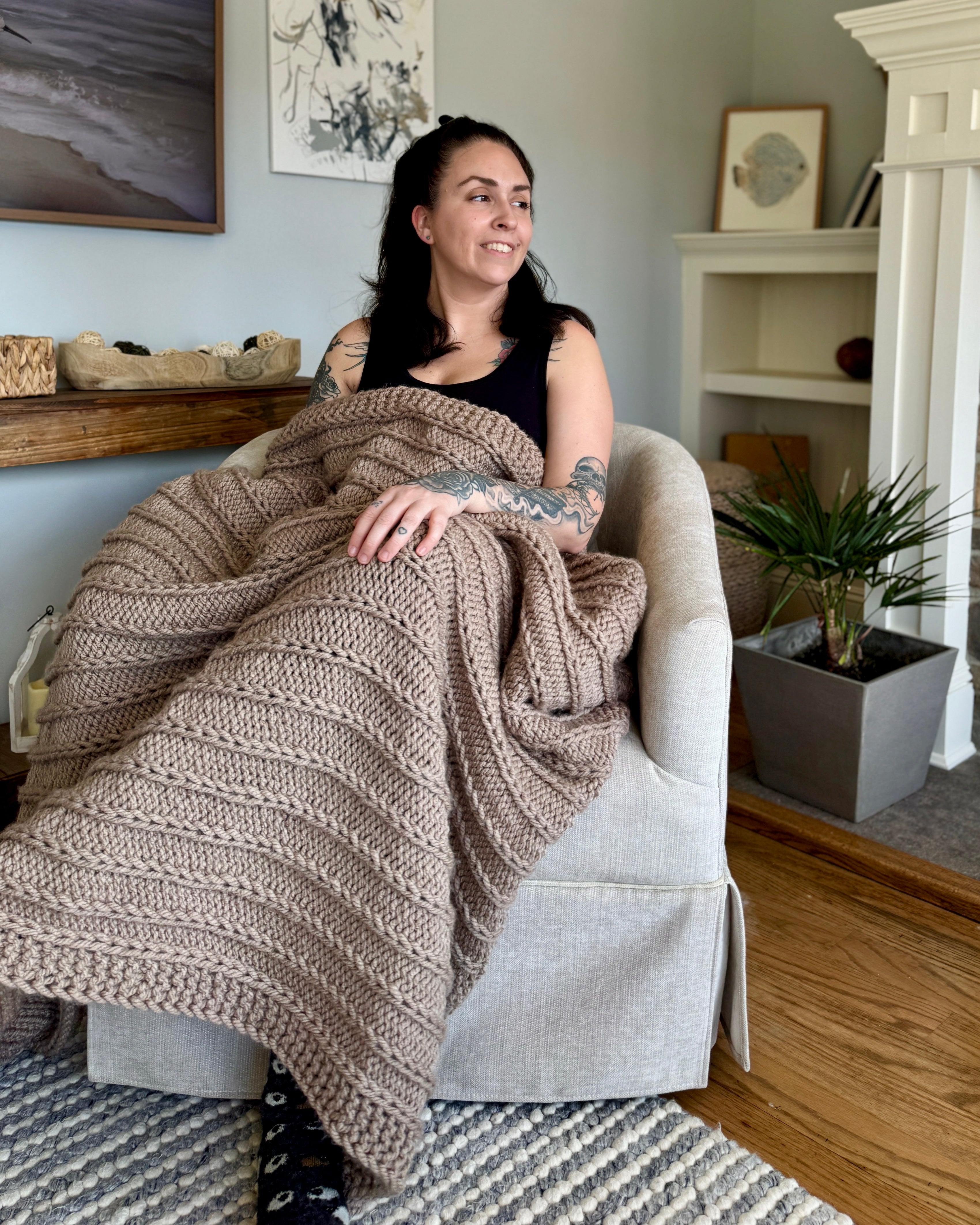 Woman sitting on a chair wrapped in a large knitted blanket in a cozy living room.