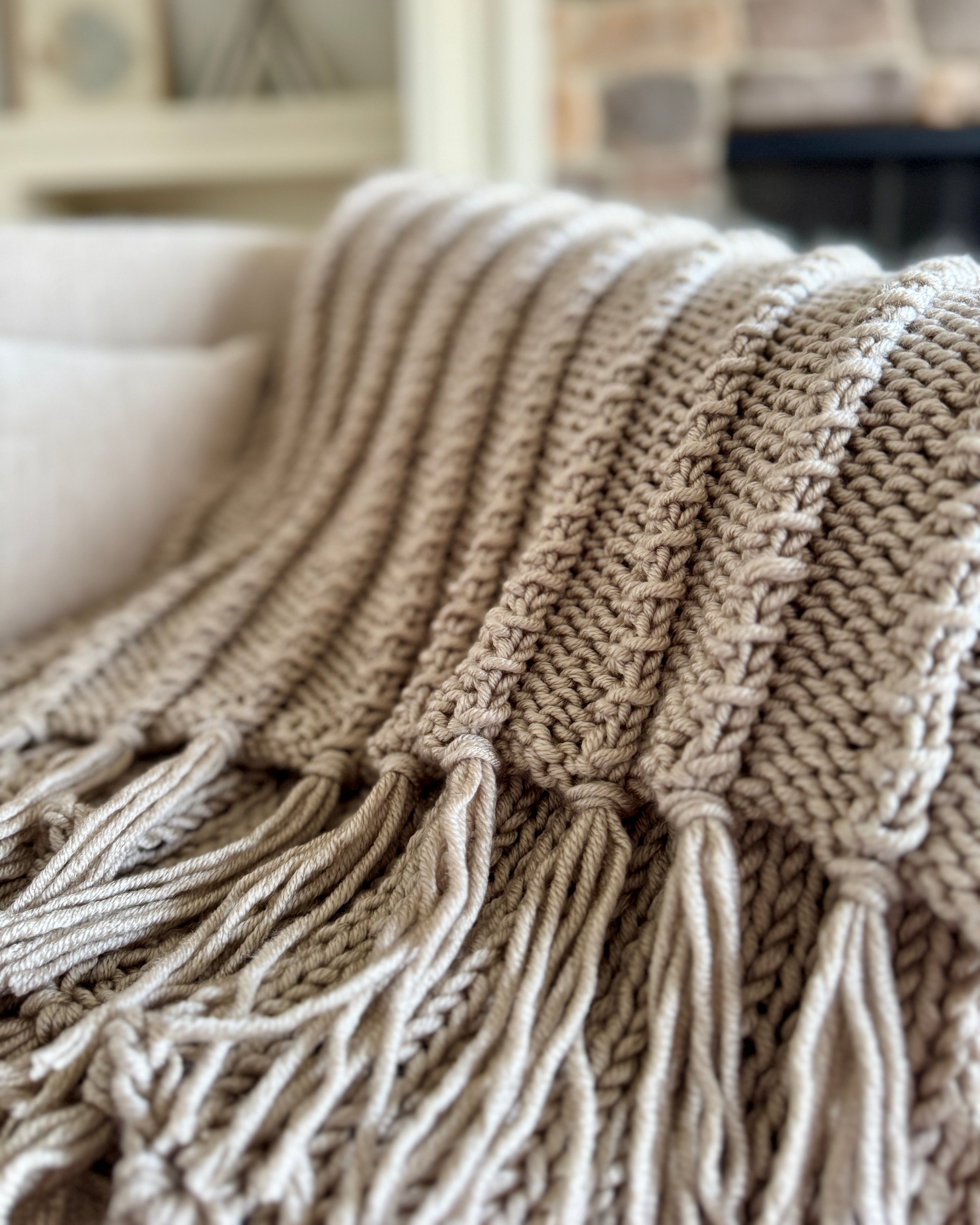 Close-up of a beige knitted blanket with tassels on a sofa.