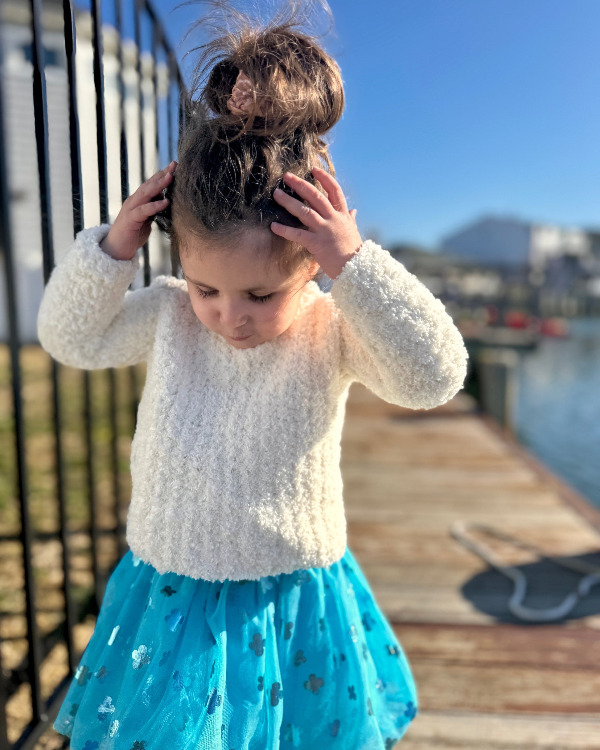 Child in a white sweater and blue skirt standing on a dock by water