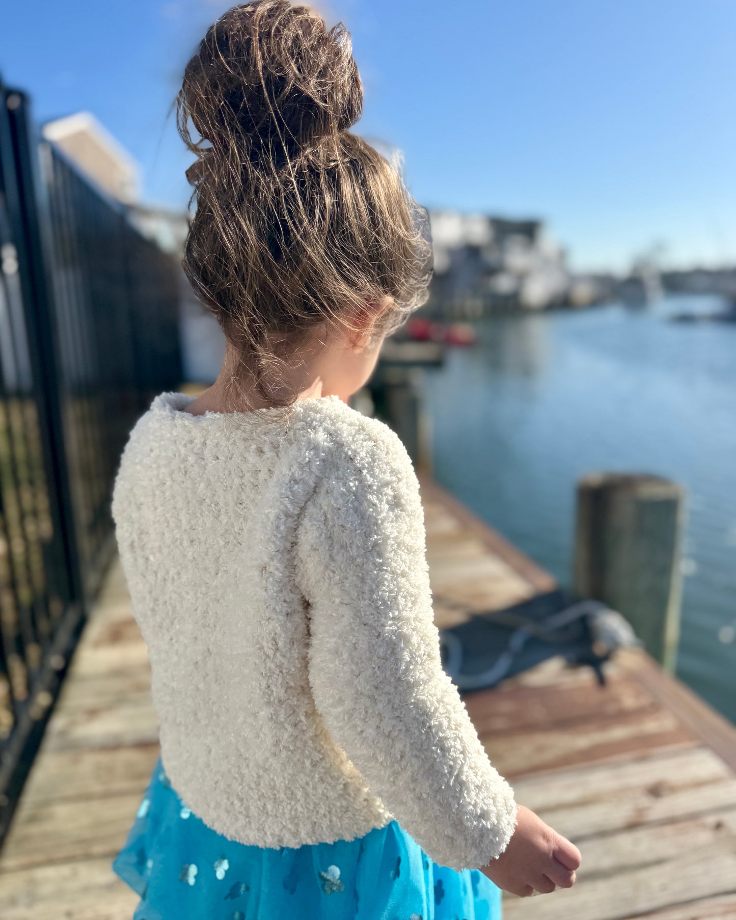 Child wearing a textured white sweater and blue dress standing on a dock by water.